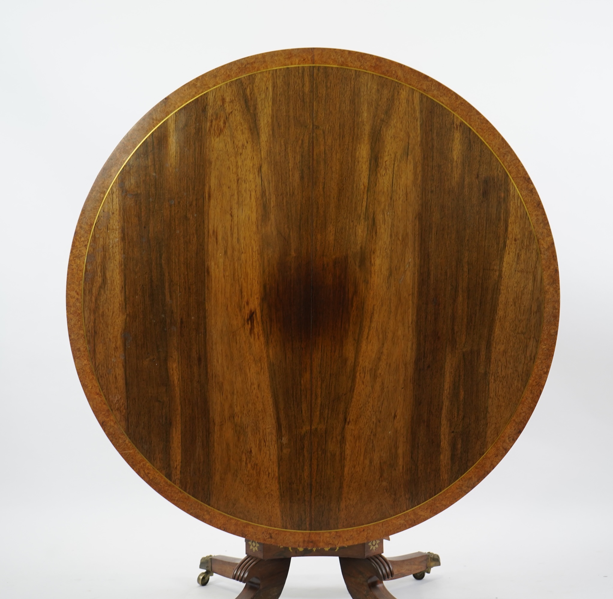A Regency cut brass inlaid and gilt metal mounted circular rosewood breakfast table, the bird's-eye maple banded tilt top on baluster column, concave platform and quadruple downswept base with cast claw caps and castors,